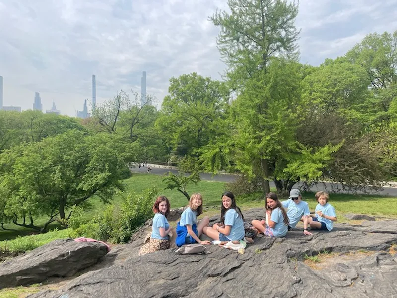 Six middle school students sitting together on a rock outcrop in Central Park with the New York City skyline visible through the trees behind them