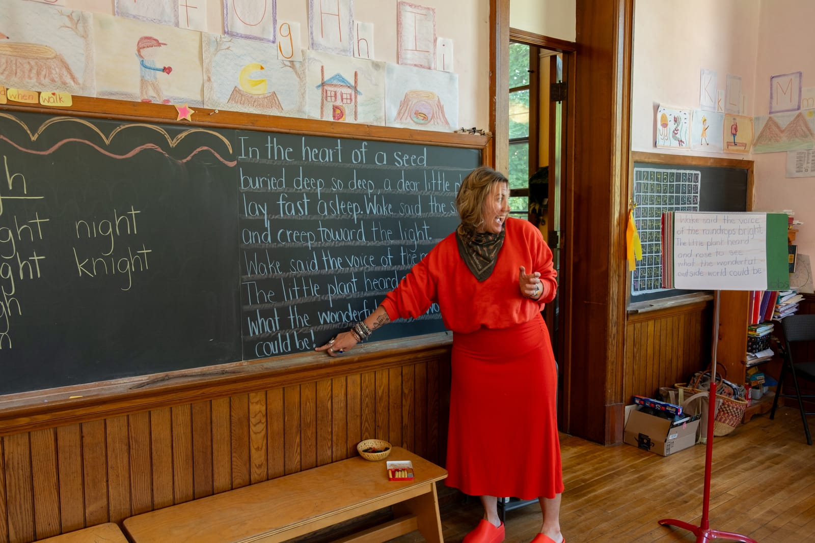 A Lower School teacher in a red dress reading a chalkboard verse aloud beneath a hand-drawn classroom alphabet