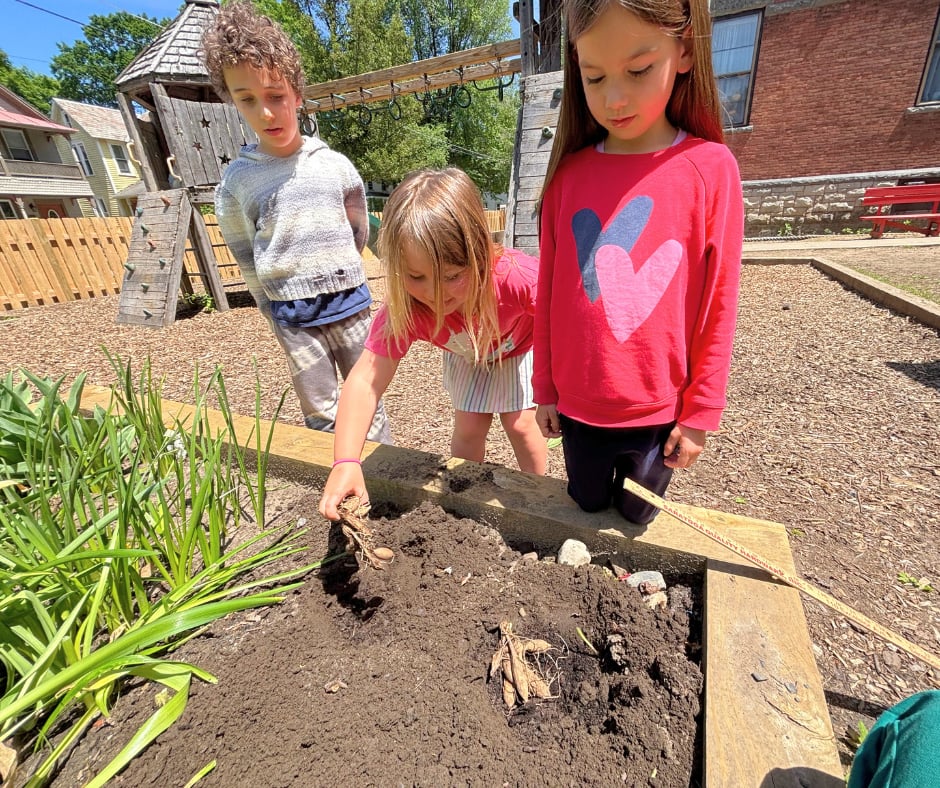 Three children leaning over a raised garden bed on the playground, working the dark soil with their hands