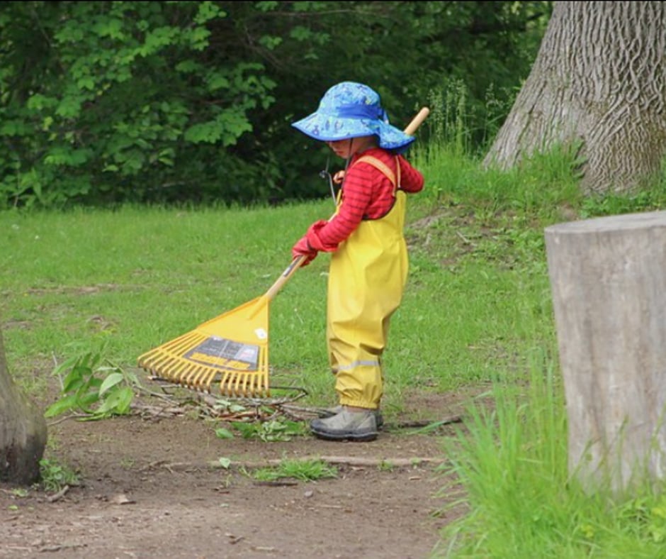 A young child in yellow rain overalls and a sun hat raking sticks and leaves along a woodland path