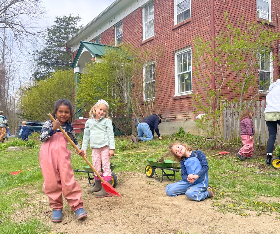 Young children with rakes and wheelbarrows working together on a spring cleanup in the yard outside the brick schoolhouse