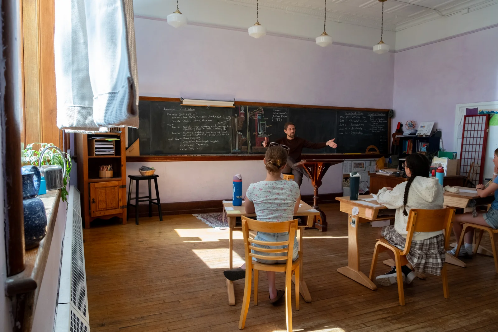 A grades-class teacher gesturing toward a chalkboard filled with American Civil War lesson notes as students sit at wooden desks in a sunlit classroom