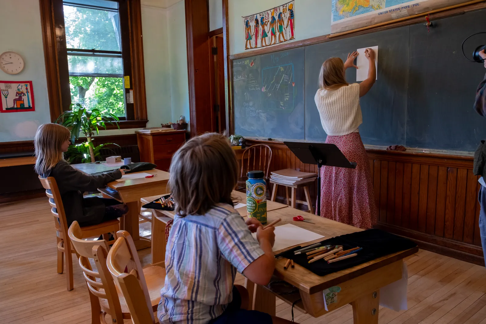 A teacher in a cream sweater pins a paper to the chalkboard at the front of a sunlit, wood-paneled Waldorf classroom while two children work with colored pencils at wooden desks near tall windows