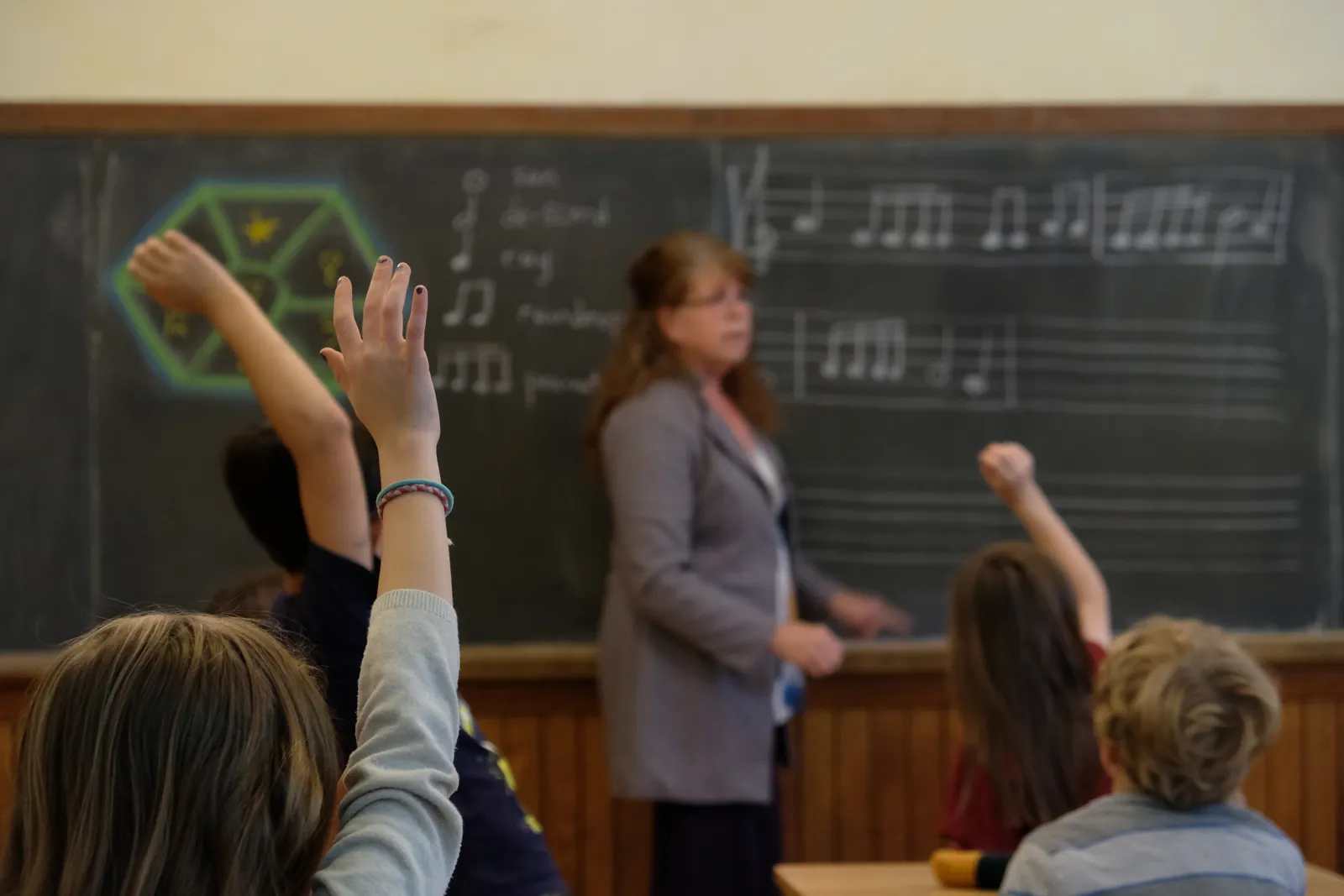 Students raising their hands in a Waldorf classroom as a teacher leads a music lesson at a chalkboard with hand-drawn notation