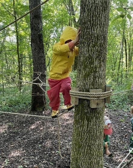 A child in a yellow jacket climbing a rope strung between trees in the woods