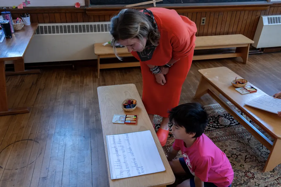 A teacher leans over a student’s main lesson book, reading a hand-lettered story about a little plant rising to see the outside world.